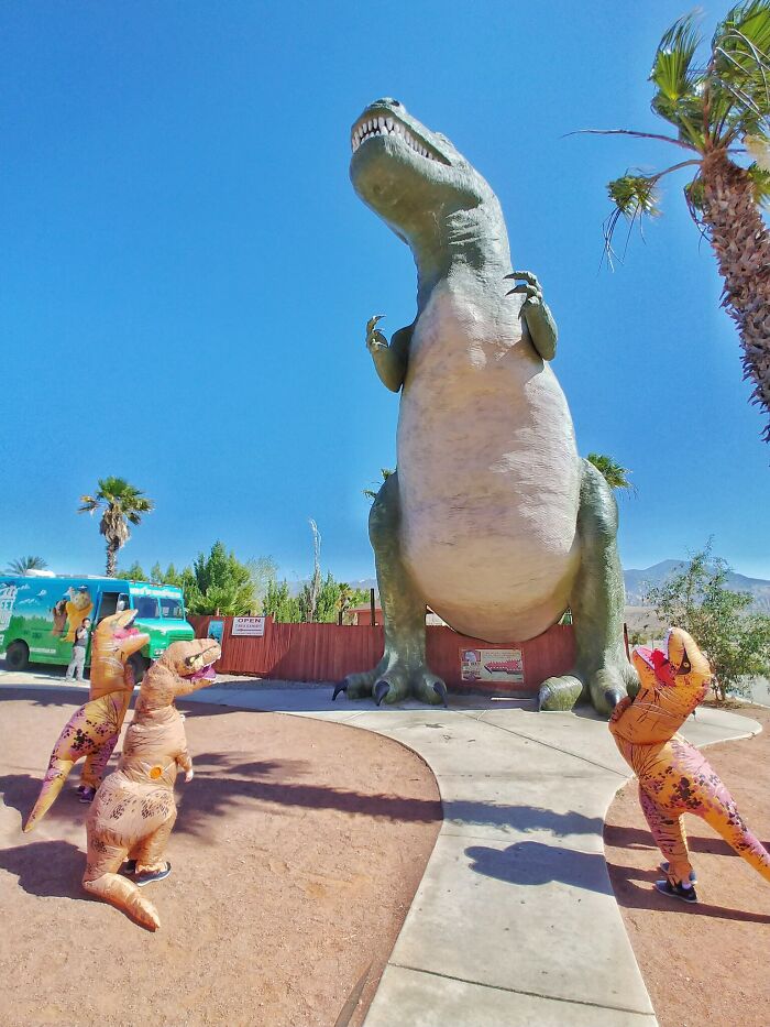 Personas con disfraces de dinosaurio frente a una estatua gigante en divertido ambiente de vacaciones de verano.