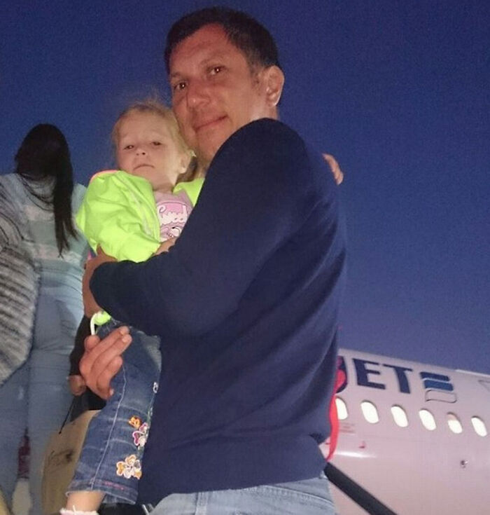 Hombre sosteniendo niña frente a avión al anochecer, foto tomada segundos antes de un desastre impactante.