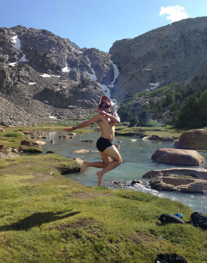 Hombre saltando divertido en un paisaje natural junto a río y montañas, foto de vacaciones de verano inolvidables.