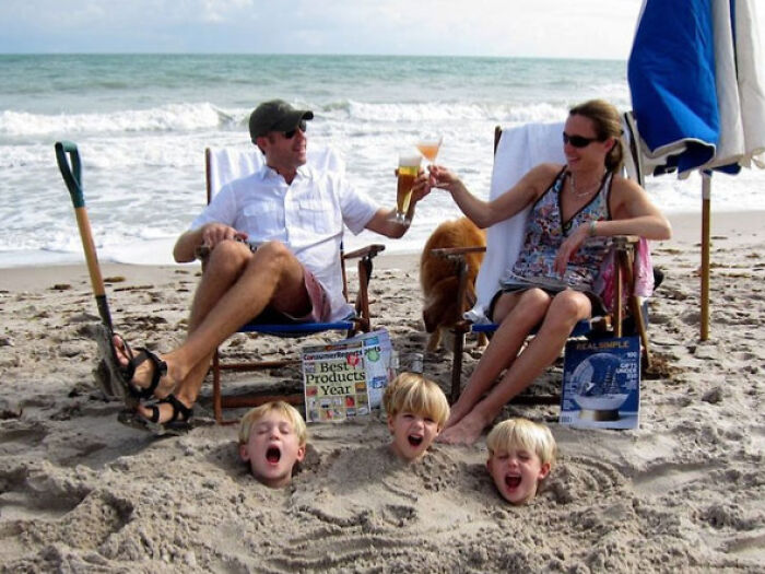 Familia disfrutando vacaciones de verano en la playa con niños enterrados en la arena y brindis relajado junto al mar.