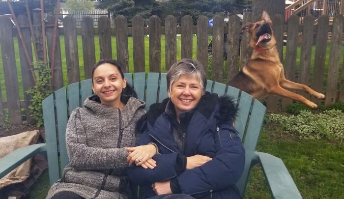 Dos mujeres sonrientes sentadas en un banco al aire libre mientras un perro salta arruinando la foto perfecta de mascotas.