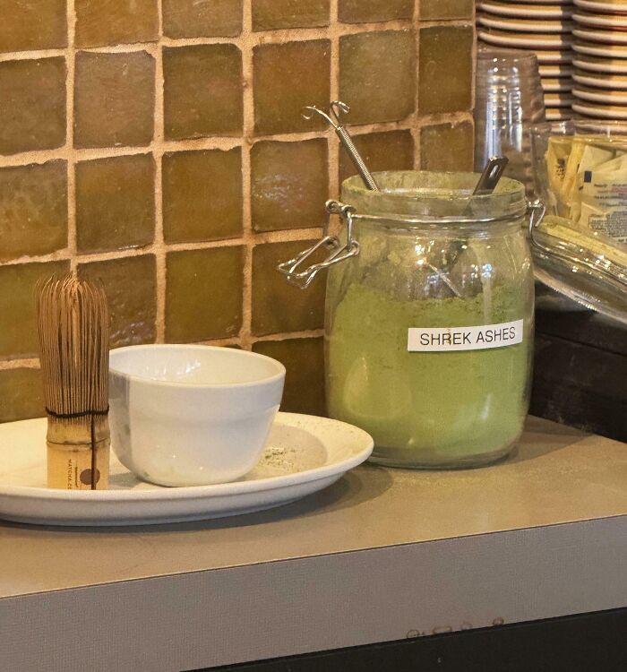 Frasco con polvo verde etiquetado como cenizas de Shrek, taza blanca y batidor de té, ejemplo de humor ingenioso.