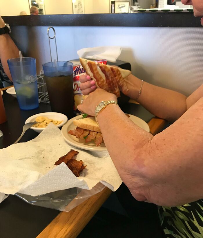 Persona en restaurante disfrutando comida y bebida, mostrando humor ingenioso en experiencias cotidianas con sándwich y bebida.