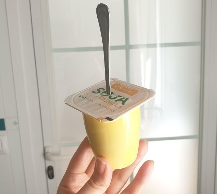 Mano sosteniendo yogur de soja con tapa original y cuchara insertada mostrando un life hack genial y práctico.