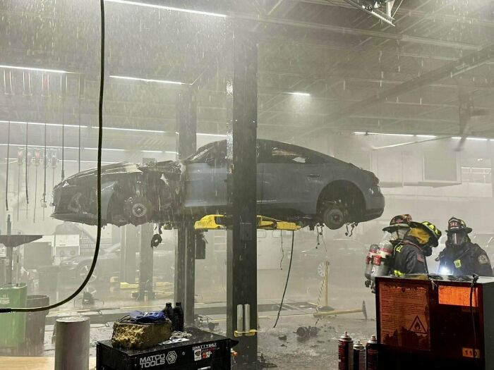 Coche levantado en taller de mecánicos con daños visibles y bomberos en escena, demostrando una situación inusual.