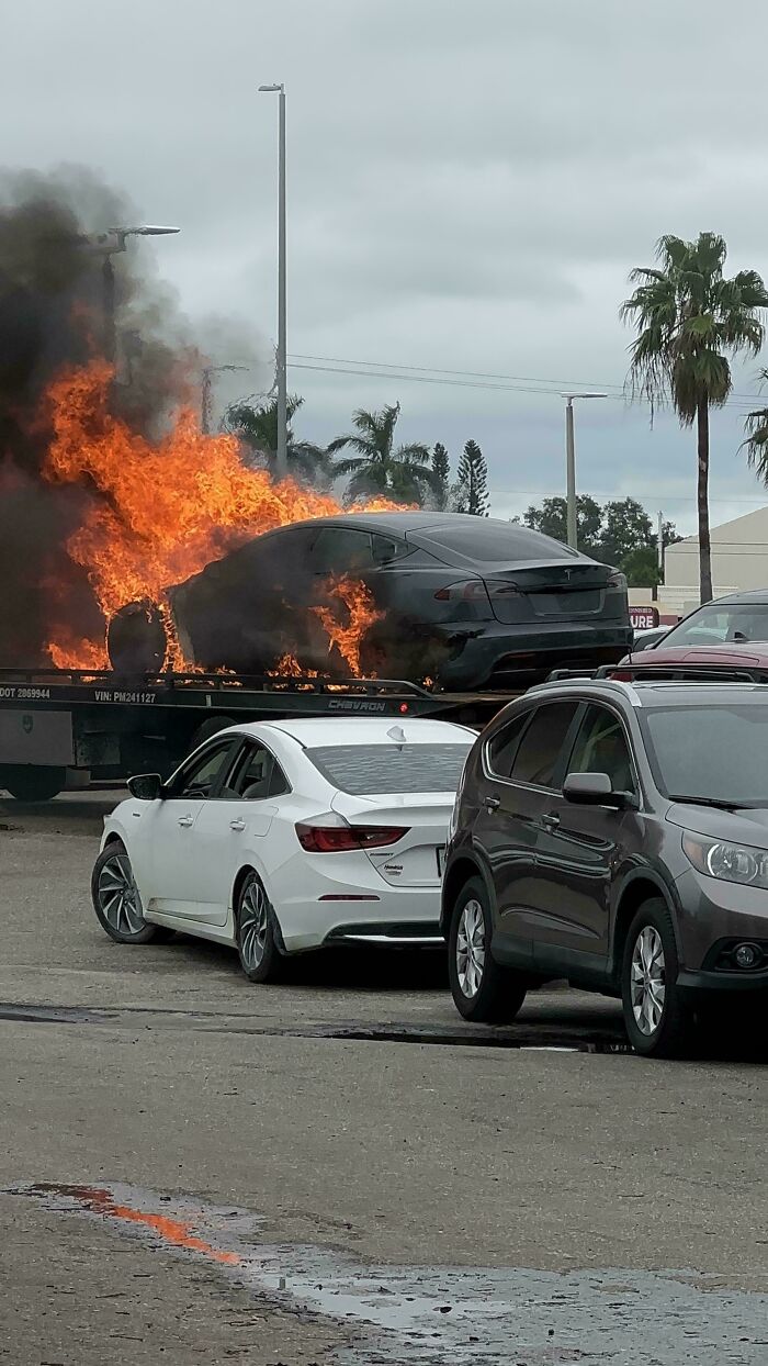 Coche en llamas en plataforma con otros vehículos cerca, mecánicos tomando fotos para demostrar el incidente automotriz.