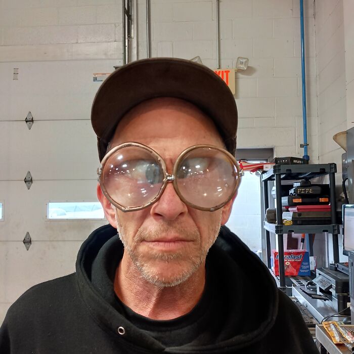 Mecánico de autos con lentes grandes en taller, mostrando una herramienta o pieza para que otros crean lo que ve.