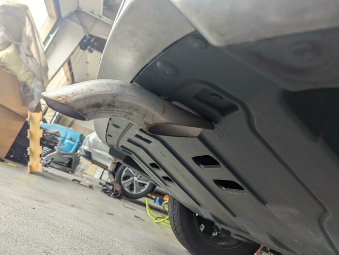 Escape de automóvil modificado y oxidado visto desde abajo en taller mecánico, imagen para mecánicos de autos que documentan reparaciones.