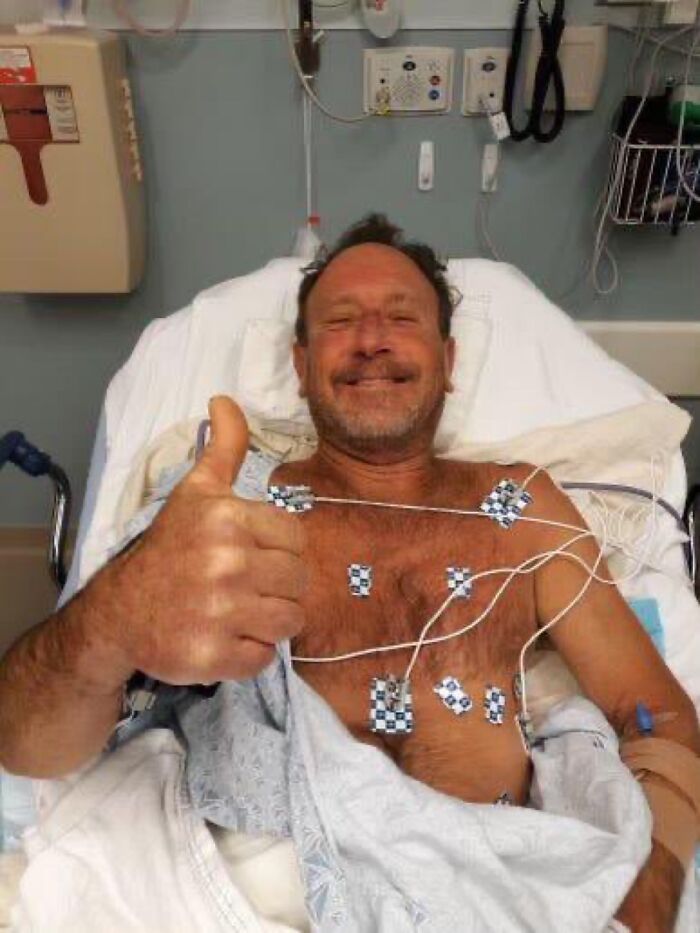 Hombre sonriente en hospital con electrodos en el pecho, dando pulgar arriba después de prueba cardíaca.