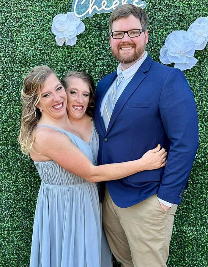 Hermanas siamesas Abby y Brittany Hensel sonríen junto a un hombre frente a fondo verde floral en evento social.