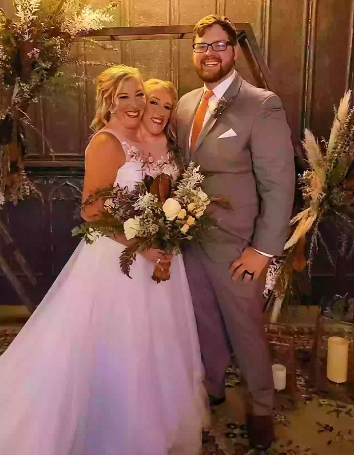 Siamesas Abby y Brittany Hensel en vestido de novia junto a hombre, con ramo floral, en interior elegante.