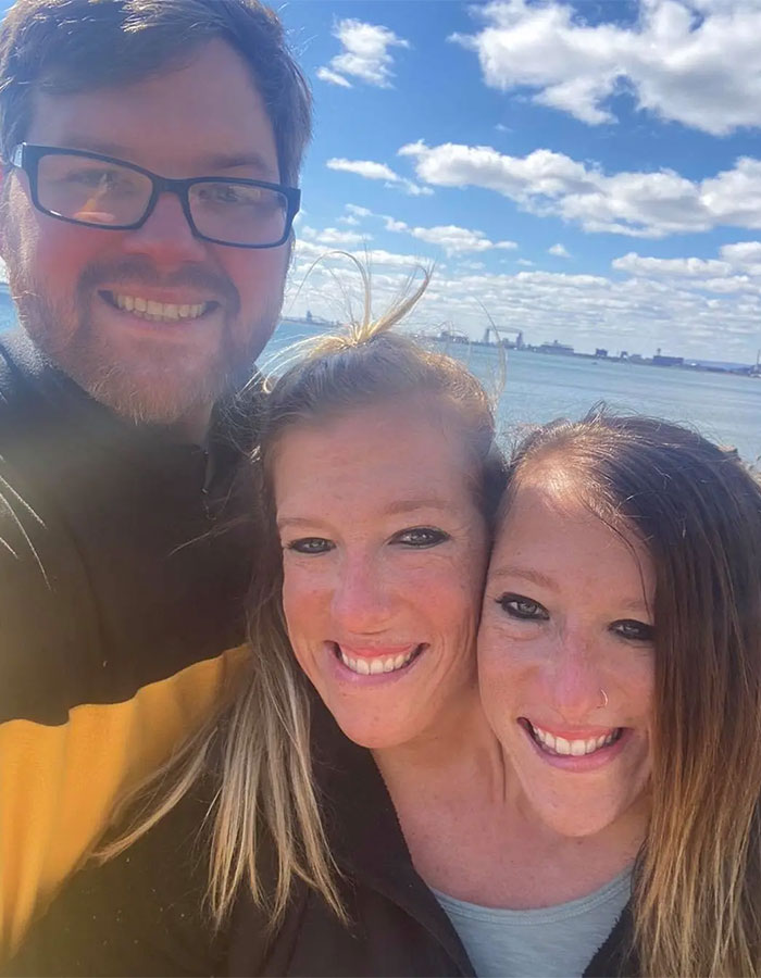 Las siamesas Abby y Brittany Hensel sonrientes al aire libre con un hombre junto al agua en un día soleado.