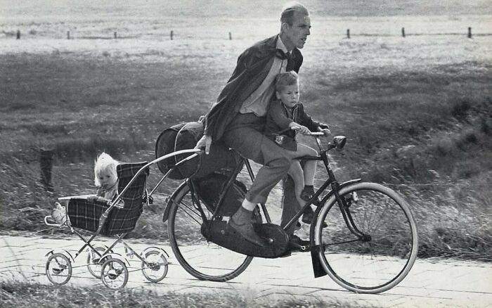 Hombre en bicicleta con dos niños, una en asiento delantero y otro en cochecito, en foto antigua y contexto histórico.
