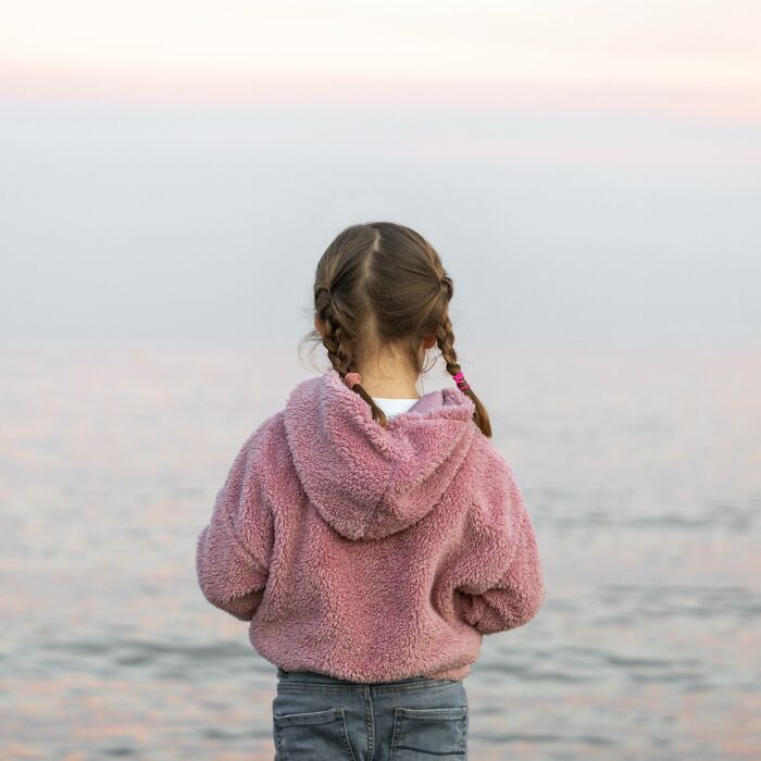 Niña con chaqueta rosa de espaldas mirando al mar al atardecer, ilustrando el contenido de Reddit para valientes.