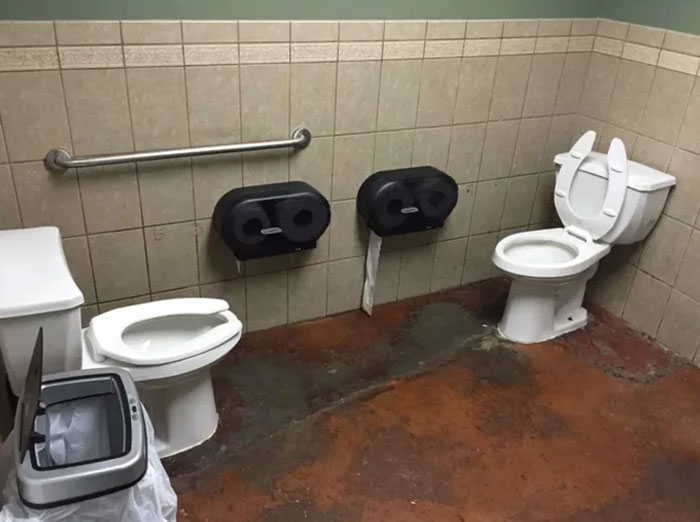 Baño con dos inodoros juntos y dispensadores de papel en medio, mostrando uno de los fallos de diseño más divertidos.