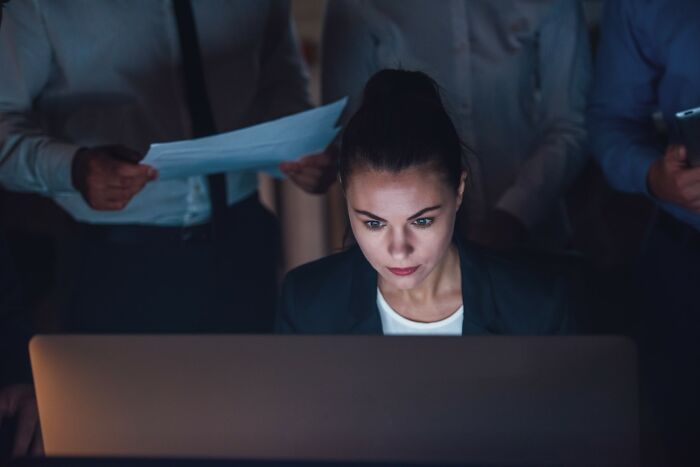 Detective privado concentrada trabajando de noche en computadora, con colegas revisando documentos en oficina oscura.