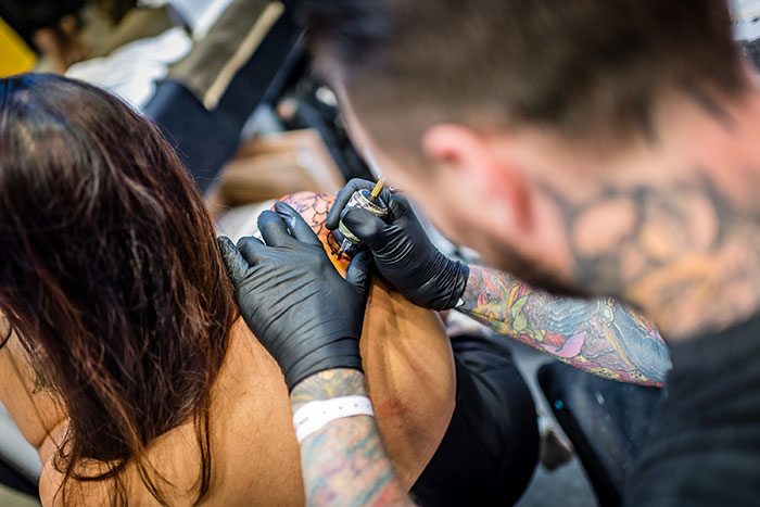Tatuador con guantes negros realizando un tatuaje en el brazo de una mujer, mostrando aspectos inquietantes de profesiones.