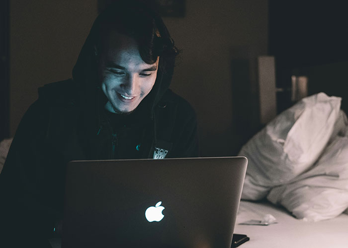 Joven usando laptop en cuarto oscuro aprovechando vacíos legales para explotar oportunidades antes de ser descubierto.