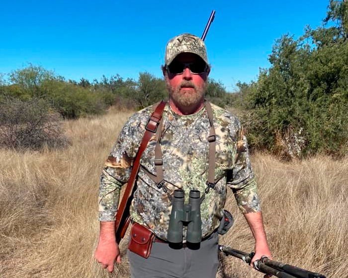 Cazador millonario de trofeos en camuflaje con rifle y binoculares en safari entre hierba alta y cielo azul.