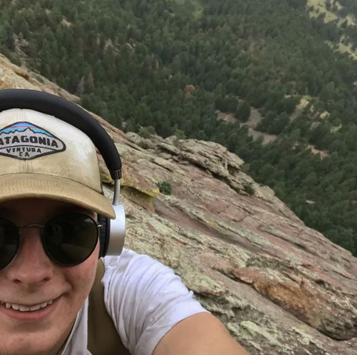 Joven con gorra y audífonos tomando selfie en un acantilado, momentos antes de un desastre inesperado.