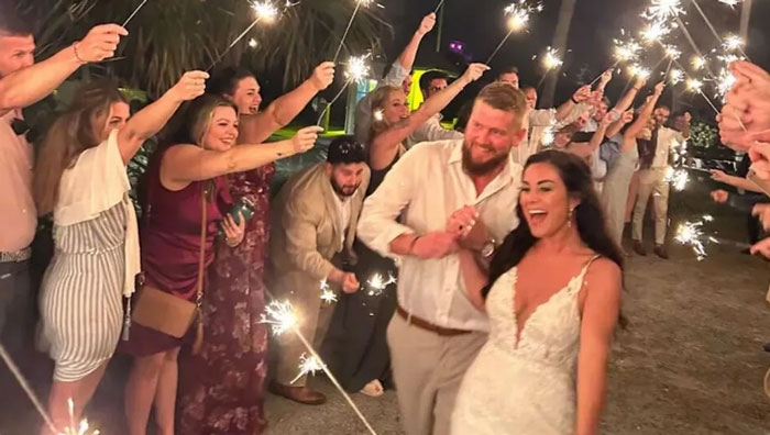 Pareja feliz en boda rodeada de invitados con bengalas, momentos capturados segundos antes de un desastre inesperado.