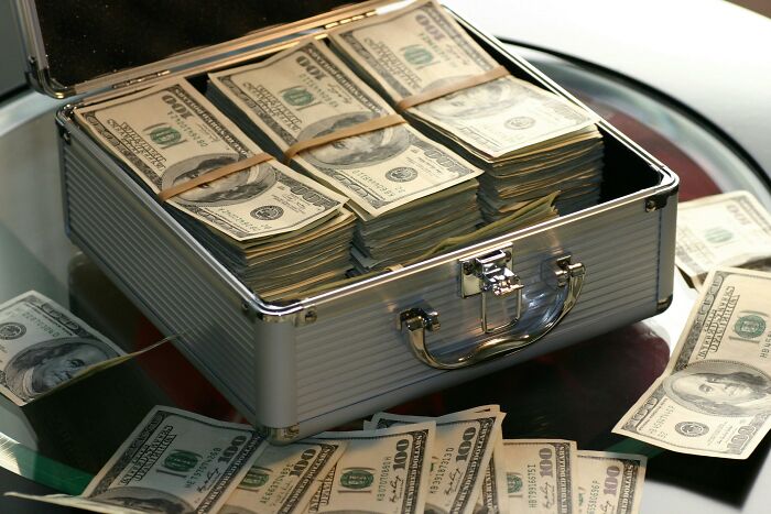 Maletín abierto con fajos de billetes de 100 dólares y billetes sueltos sobre una superficie reflectante.