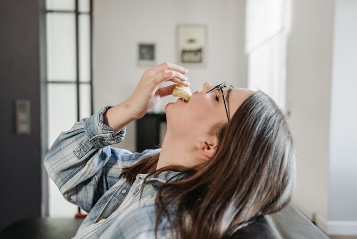 Mujer con gafas compartiendo un pequeño secreto mientras come un pastelillo en casa de forma relajada.