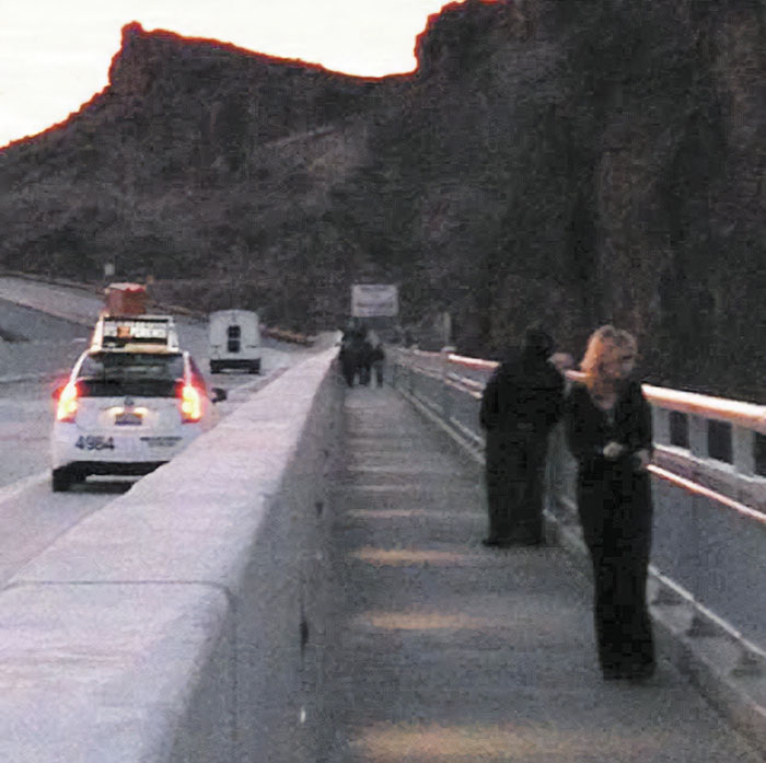 Persona caminando en puente acompañado de policía, foto tomada segundos antes de un desastre impactante en carretera y montaña.