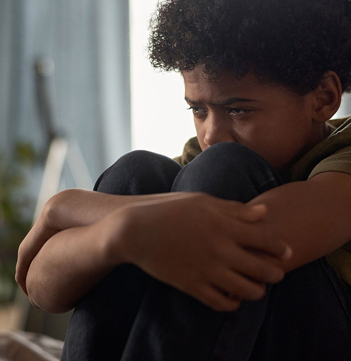 Niño triste abrazando sus rodillas en casa, ilustrando experiencias aterradoras de personas que crecieron con un psicópata.