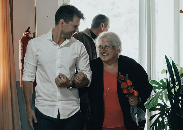 Hombre joven sonriendo junto a suegra mayor que sostiene flores en ambiente interior, mostrando relación entre personas y suegras.