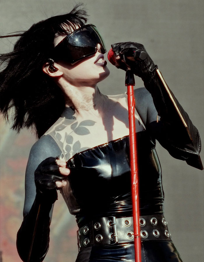 Cantante con tatuajes negros en el 80% del cuerpo, usando gafas oscuras y guantes, sostiene un micrófono rojo en el escenario.