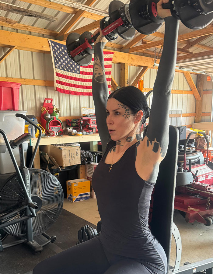Kat Von D levantando pesas en gimnasio, mostrando su tatuaje que cubre gran parte de su cuerpo de negro.