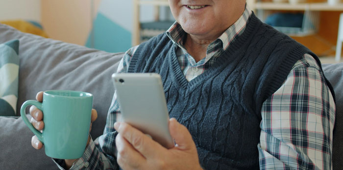 Hombre mayor sentado en sofá usando teléfono móvil mientras sostiene una taza, ilustrando cómo los teléfonos revelan verdades ocultas. Hombre mayor sentado en sofá usando teléfono móvil mientras sostiene una taza, ilustrando cómo los teléfonos revelan verdades ocultas.
