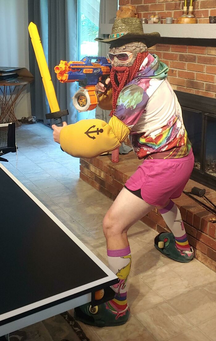 Persona con uniforme hilarante usando sombrero, gafas, camisa colorida, guantes inflables y sosteniendo una espada y pistola de juguete.