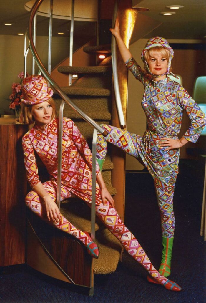 Dos mujeres con uniformes coloridos y estampados extravagantes posando en una escalera en un interior retro.