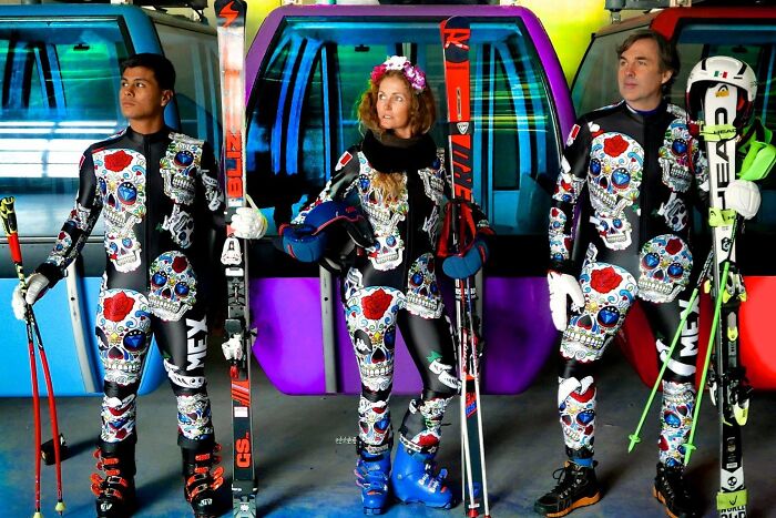 Tres esquiadores con uniformes hilarantes de cráneos decorados, puestos frente a góndolas coloridas en estación de esquí.