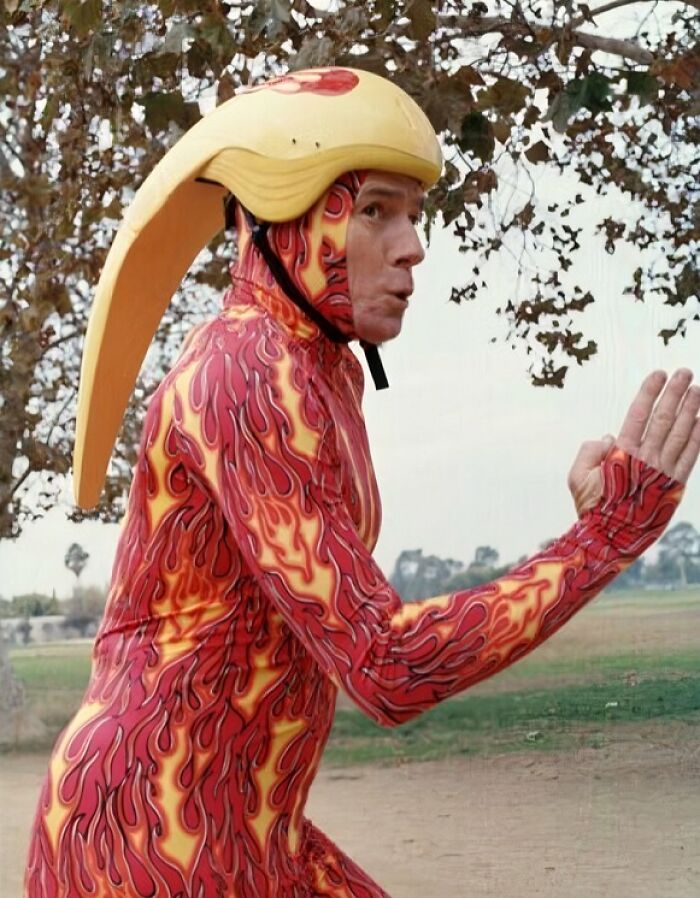 Hombre con uniforme hilarante de llamas y casco amarillo en parque, diseño que hace cuestionar su origen creativo.