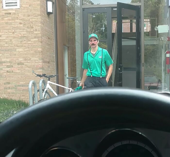 Hombre con uniforme verde y tirantes llamativos sosteniendo gorra frente a una bicicleta junto a una entrada de edificio.