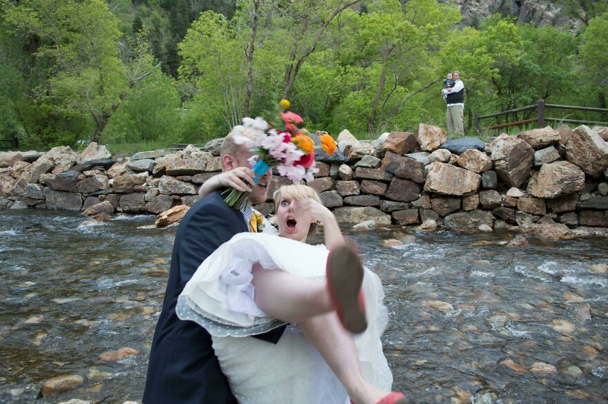 Novio levantando a su esposa cerca del arroyo tras la boda, escena peligrosa y divertida con flores y vestido blanco.