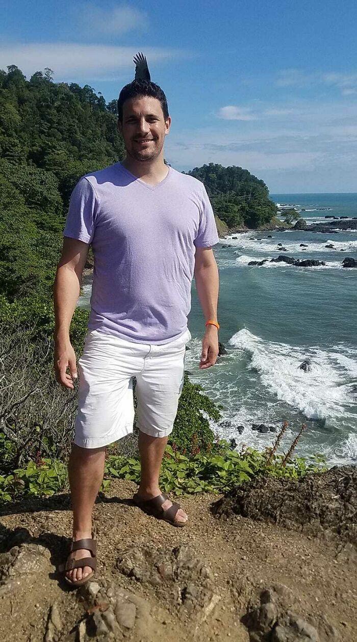 Hombre sonriente en la playa con un pájaro posado que crea una foto mejor de lo esperado con naturaleza al fondo.