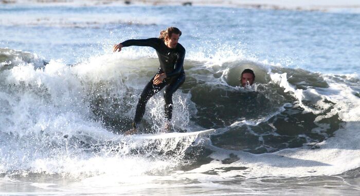 Persona surfeando una ola grande mientras otra persona aparece inesperadamente en la foto, capturando una imagen mejor.