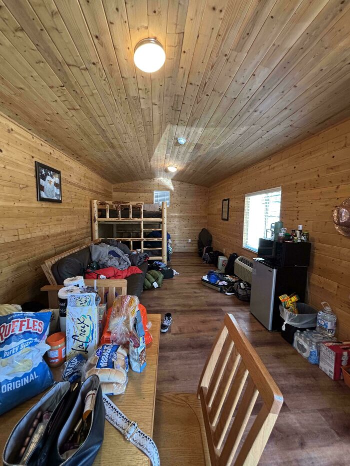 Interior de cabaña de madera con litera, mesa con comida y ropa amontonada, foto de vacaciones inesperada.