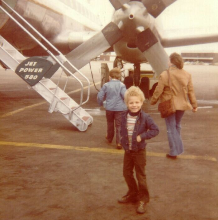 Niño sonriente con chaqueta azul posando frente a un avión antiguo mientras otros caminan en el aeropuerto vintage. Niño sonriente con chaqueta azul posando frente a un avión antiguo mientras otros caminan en el aeropuerto vintage.