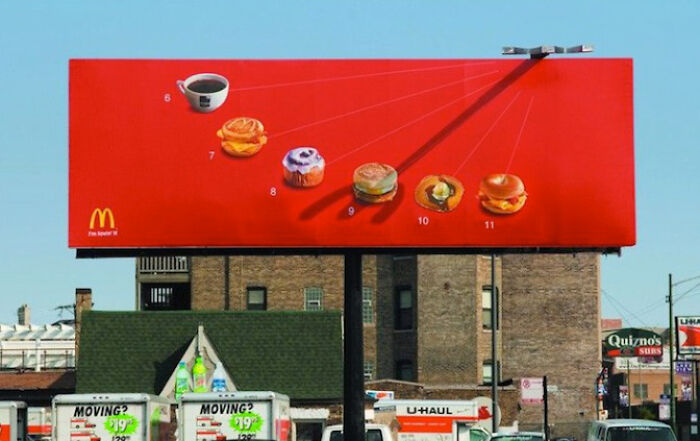 Cartel publicitario divertido y creativo de McDonald's con café y diversos alimentos sobre fondo rojo captado en ciudad urbana.