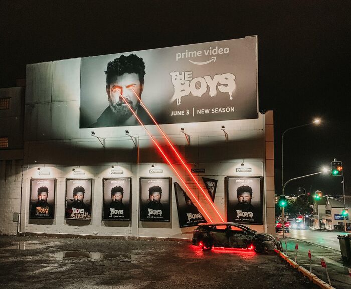 Valla publicitaria divertida y extraña de The Boys con láseres rojos saliendo de los ojos al coche estacionado frente a ella.