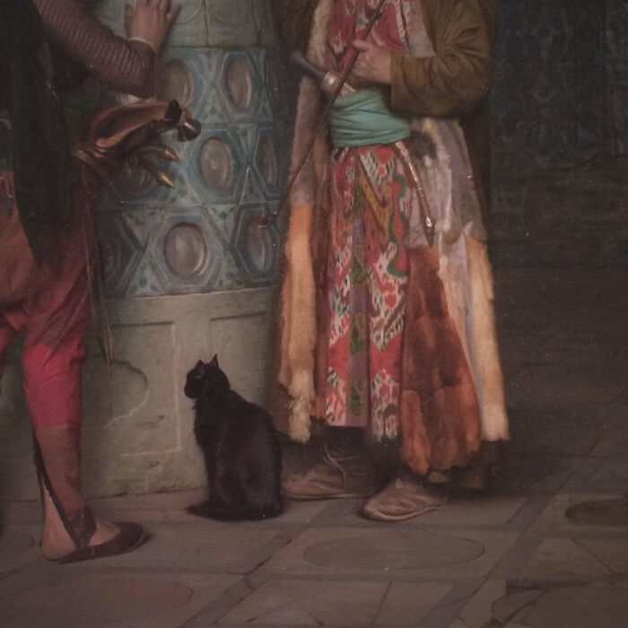 Gato medieval negro sentado junto a personas con ropa histórica en una escena humorística de imágenes medievales de gatos.