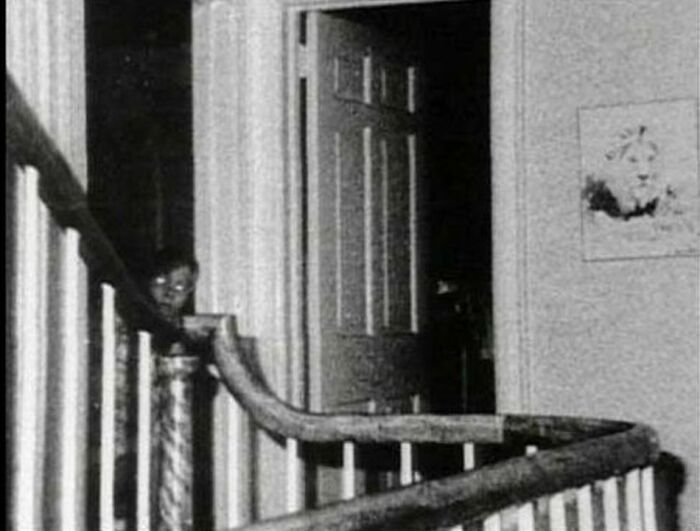 Niño con ojos brillantes parado detrás de una barandilla en una casa antigua, foto real inexplicada misteriosa.