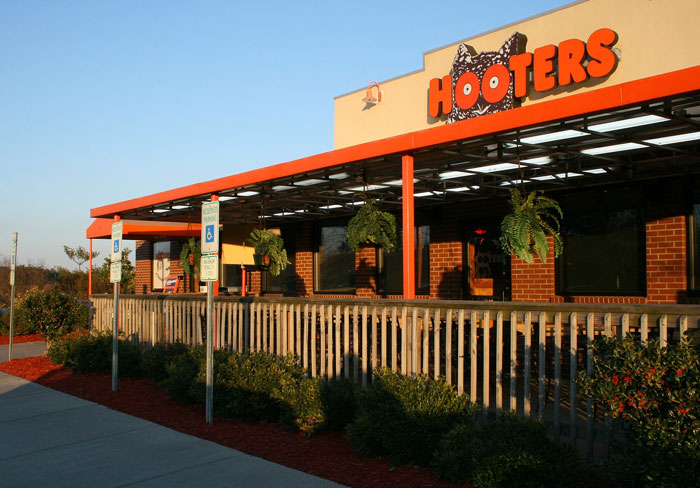 Fachada de restaurante Hooters al atardecer, con plantas colgantes y señales de estacionamiento para discapacitados. Fachada de restaurante Hooters al atardecer, con plantas colgantes y señales de estacionamiento para discapacitados.