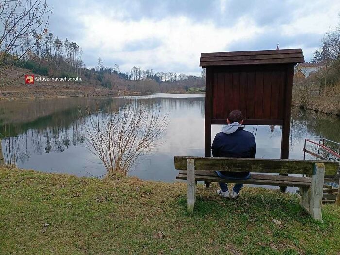 Persona sentada en un banco frente a un cartel de construcción mal ubicado junto a un lago, mostrando fallos divertidos.