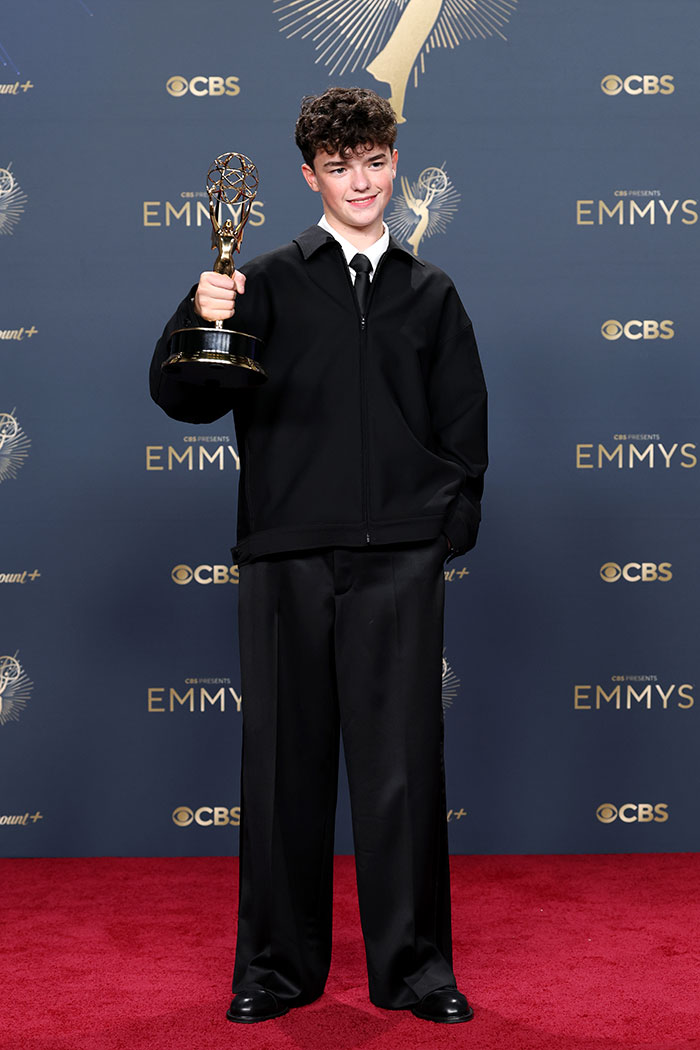 Joven vestido con atuendo negro sostiene premio Emmy en la alfombra roja de los Emmys 2025, moda y estilo destacado.